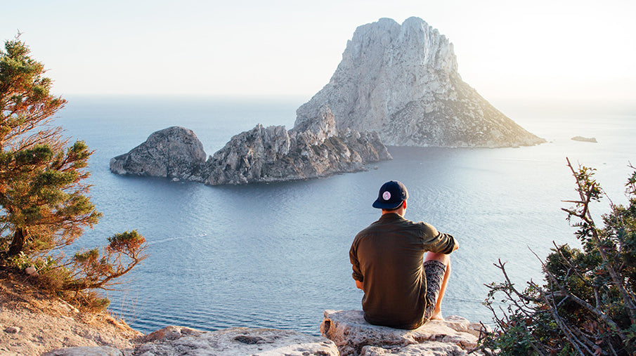 Man sitting on cliff top overlooking a bay with the sunsetting over the ocean