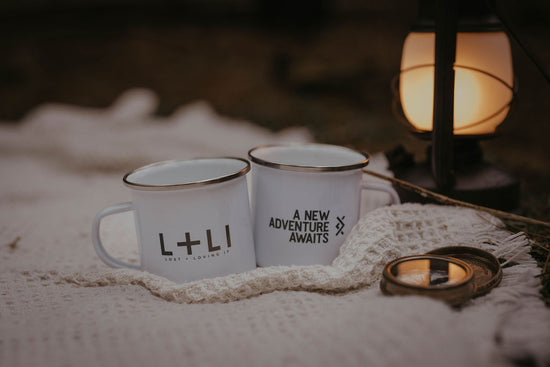 white enamel mug with 'A New Adventure Awaits' written on it and another with L+LI printed on it, on a blanket in a wood with an oil lamp and compass